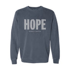 A denim blue crewneck sweatshirt featuring the word 'HOPE' printed across the chest in a white pattern of smaller words, with 'HUMBLE + HOPEFUL' written underneath.