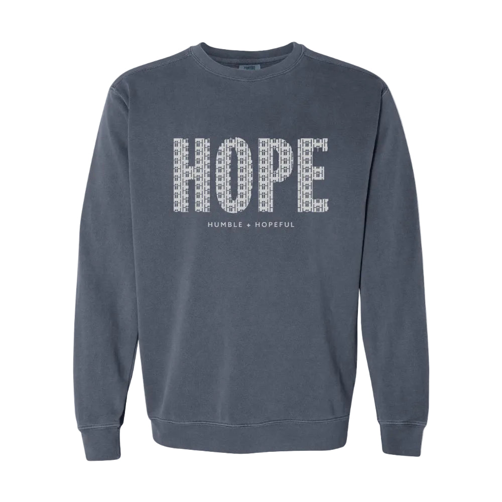 A denim blue crewneck sweatshirt featuring the word 'HOPE' printed across the chest in a white pattern of smaller words, with 'HUMBLE + HOPEFUL' written underneath.