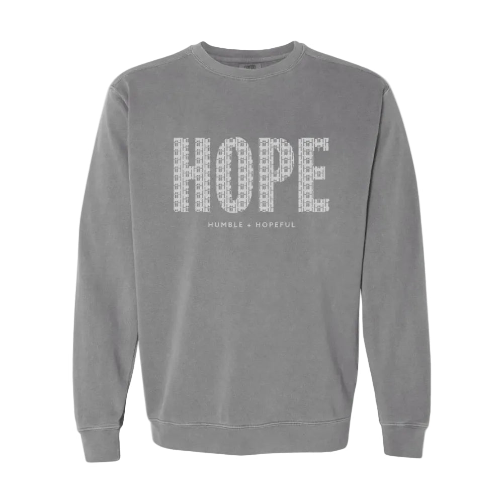 A gray crewneck sweatshirt featuring the word 'HOPE' printed across the chest in a white patterned font, with 'HUMBLE + HOPEFUL' written underneath in smaller text.