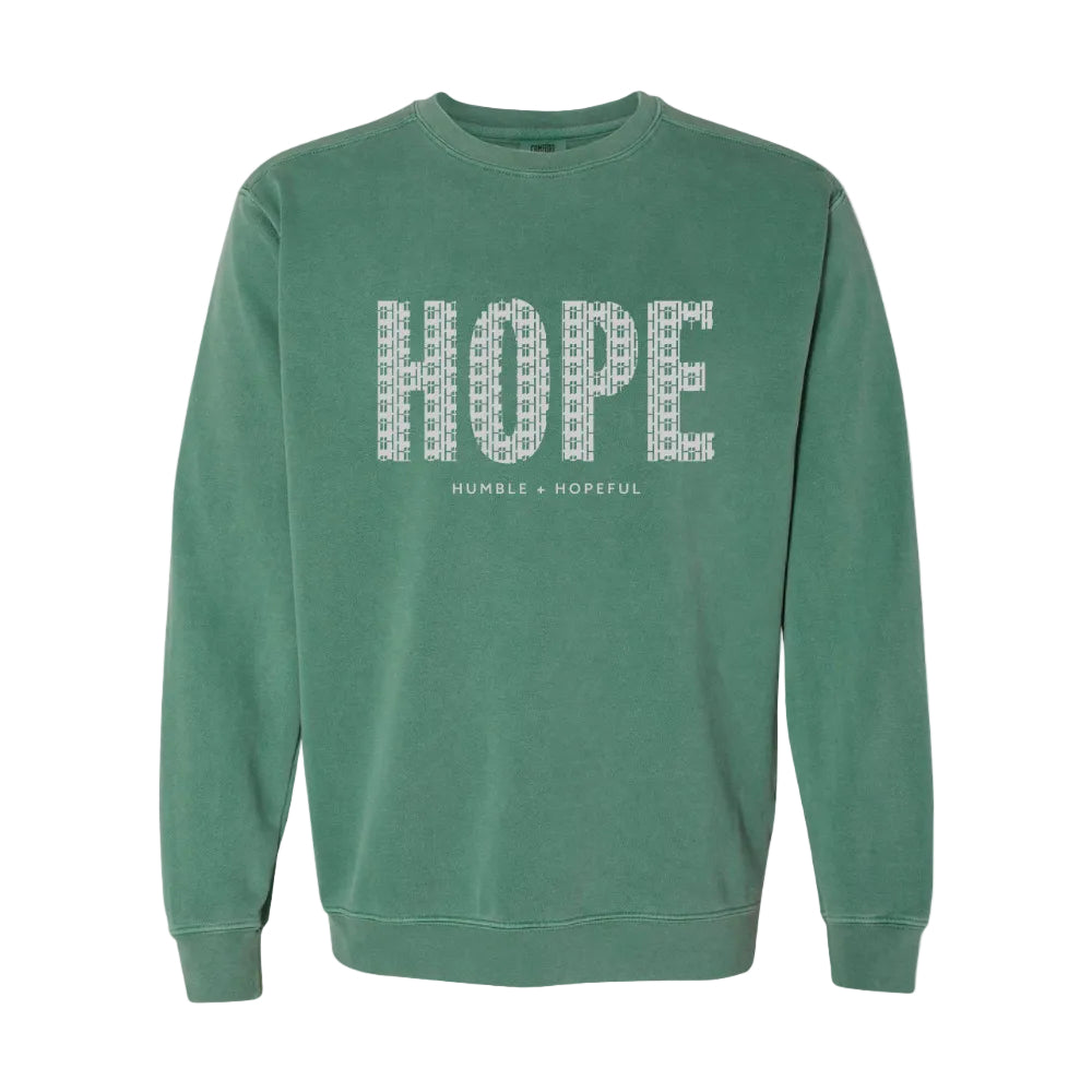 A light green crewneck sweatshirt featuring the word 'HOPE' printed across the chest in a white patterned font, with 'HUMBLE + HOPEFUL' written underneath in smaller white text.