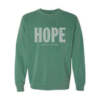 A light green crewneck sweatshirt featuring the word 'HOPE' printed across the chest in a white patterned font, with 'HUMBLE + HOPEFUL' written underneath in smaller white text.