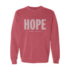 A crimson crewneck sweatshirt featuring the word 'HOPE' printed across the chest in a white pattern of small words, with 'HUMBLE + HOPEFUL' written underneath in smaller text.