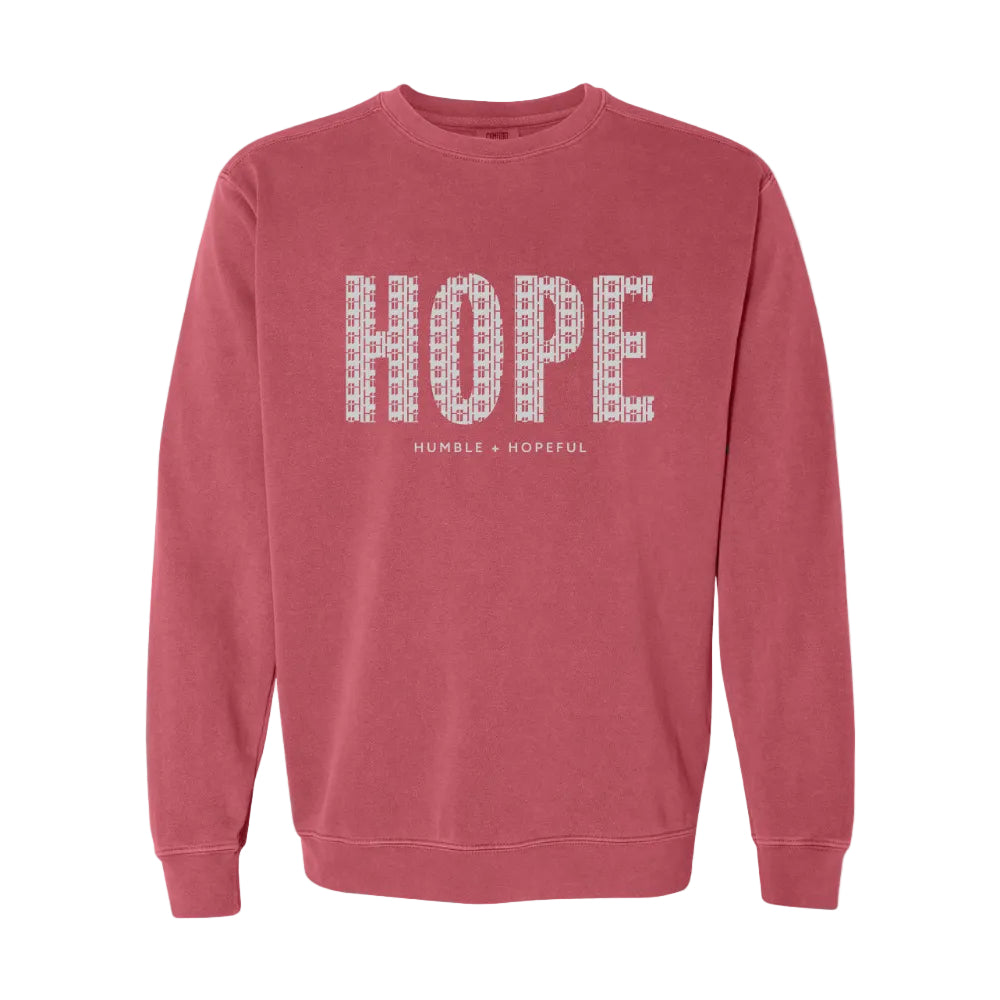 A crimson crewneck sweatshirt featuring the word 'HOPE' printed across the chest in a white pattern of small words, with 'HUMBLE + HOPEFUL' written underneath in smaller text.