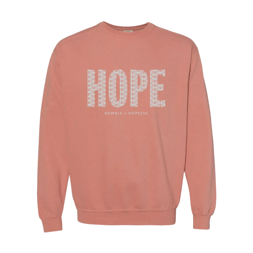 A terracotta crewneck sweatshirt featuring the word 'HOPE' printed across the chest in a white patterned font, with 'HUMBLE + HOPEFUL' written underneath in smaller text.
