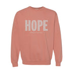 A terracotta crewneck sweatshirt featuring the word 'HOPE' printed across the chest in a white patterned font, with 'HUMBLE + HOPEFUL' written underneath in smaller text.