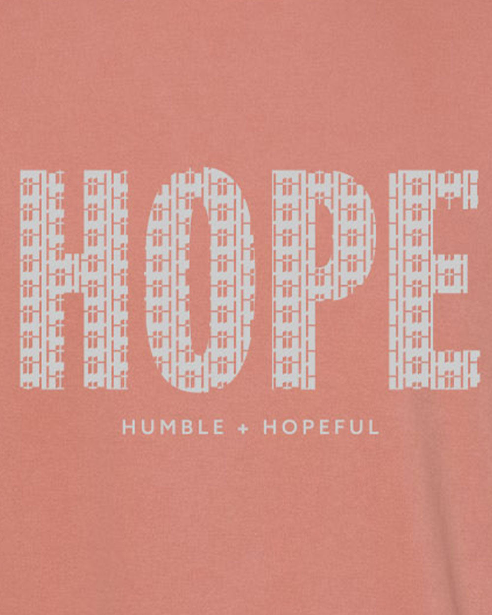 A close-up of a terracotta garment featuring a large white graphic print that spells 'HOPE' with a cross-hatch pattern inside each letter, and the phrase 'HUMBLE + HOPEFUL' printed below in smaller white text.