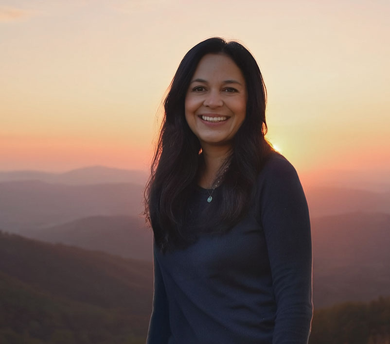 The owner of Humble and Hopeful standing outdoors against a sunset over layered mountain ridges. She is wearing a dark navy long-sleeve top and a small silver pendant necklace.