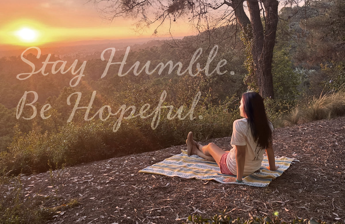 The Humble+Hopeful owner sits on a patterned blanket on a hillside at sunset, facing a scenic landscape with hills, trees, and a glowing sun. Large overlaid text reads 'Stay Humble. Be Hopeful."