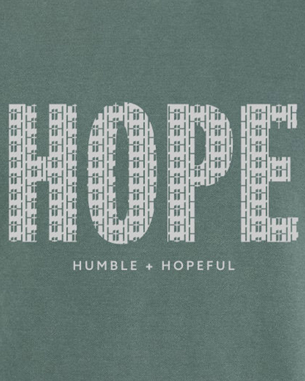A close-up of a sage green fabric surface with large white block letters spelling 'HOPE' and smaller text below reading 'HUMBLE + HOPEFUL'.