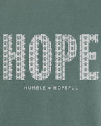 A close-up of a sage green fabric surface with large white block letters spelling 'HOPE' and smaller text below reading 'HUMBLE + HOPEFUL'.
