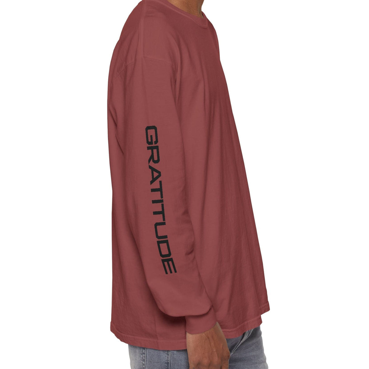 A man shown from the side wearing a brick colored long-sleeve t-shirt with 'BLESSED' printed vertically in black on the sleeve, paired with light blue jeans against a plain white background.