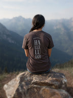 A woman sits on a large rock in a mountain landscape, viewed from behind. She is wearing a dark brown t-shirt with 'HALLELUJAH' printed vertically in white on the back.