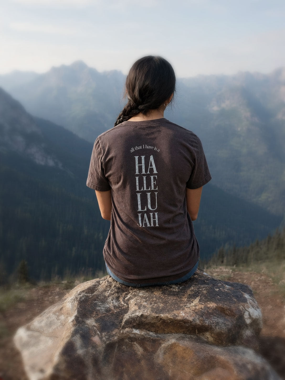 A woman sits on a large rock in a mountain landscape, viewed from behind. She is wearing a dark brown t-shirt with 'HALLELUJAH' printed vertically in white on the back.
