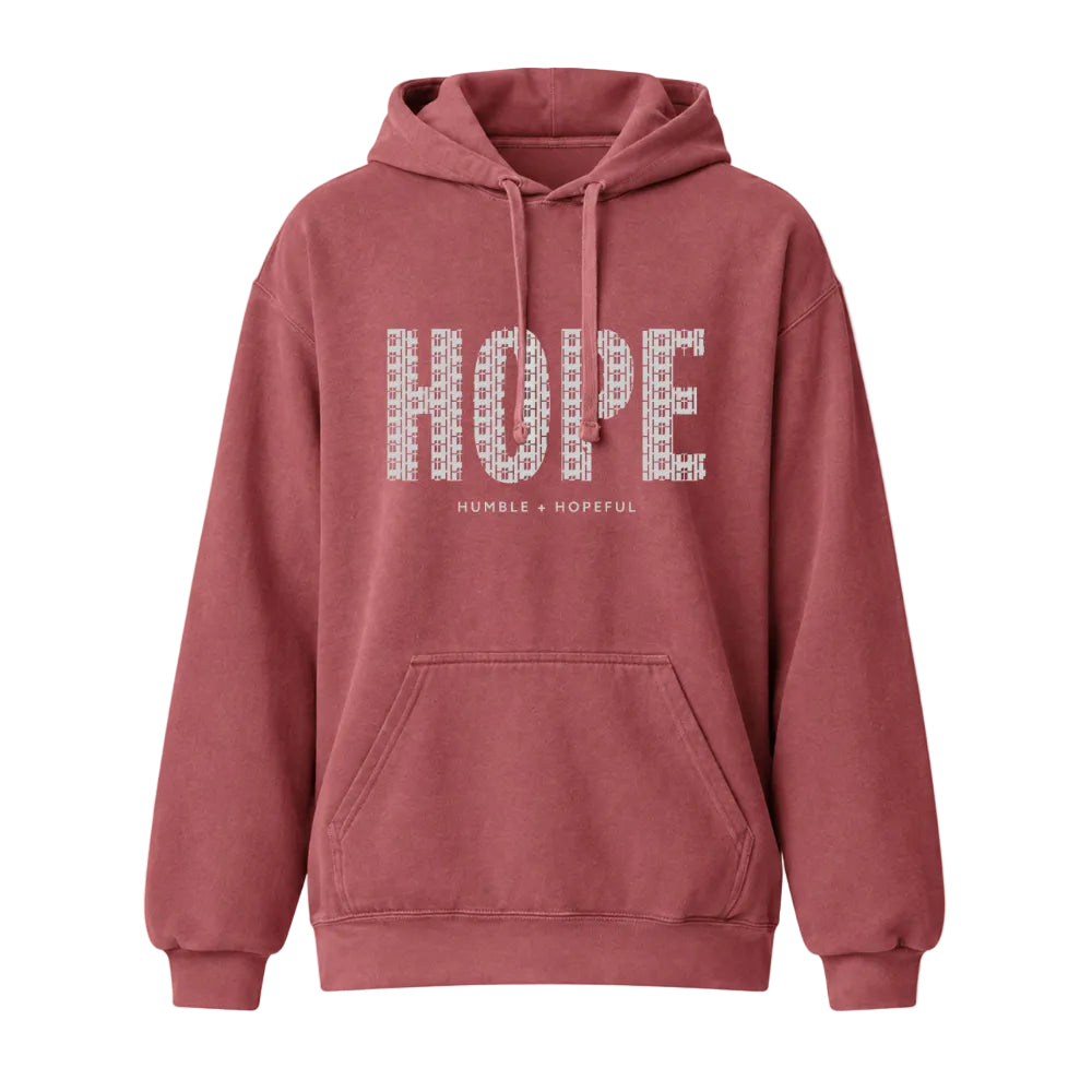 A crimson pullover hoodie with a front kangaroo pocket and drawstrings. The word 'HOPE' is printed across the chest in a white patterned font, with the words 'HUMBLE + HOPEFUL' printed underneath in a smaller font.