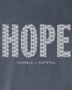 Close-up of a denim blue fabric with a large white 'HOPE' graphic made of repeating small cross-like symbols, and smaller text below reading 'HUMBLE + HOPEFUL'.