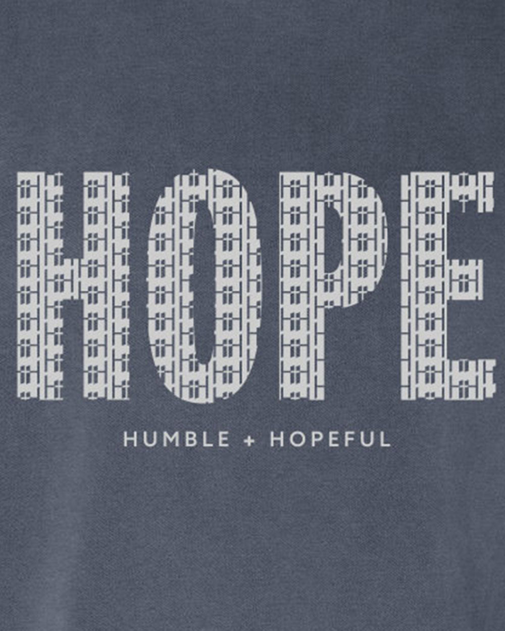 Close-up of a denim blue fabric with a large white 'HOPE' graphic made of repeating small cross-like symbols, and smaller text below reading 'HUMBLE + HOPEFUL'.