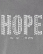 Close-up of a gray fabric surface with a large white 'HOPE' wordmark made of small cross-like shapes, and smaller text below reading 'HUMBLE + HOPEFUL'.