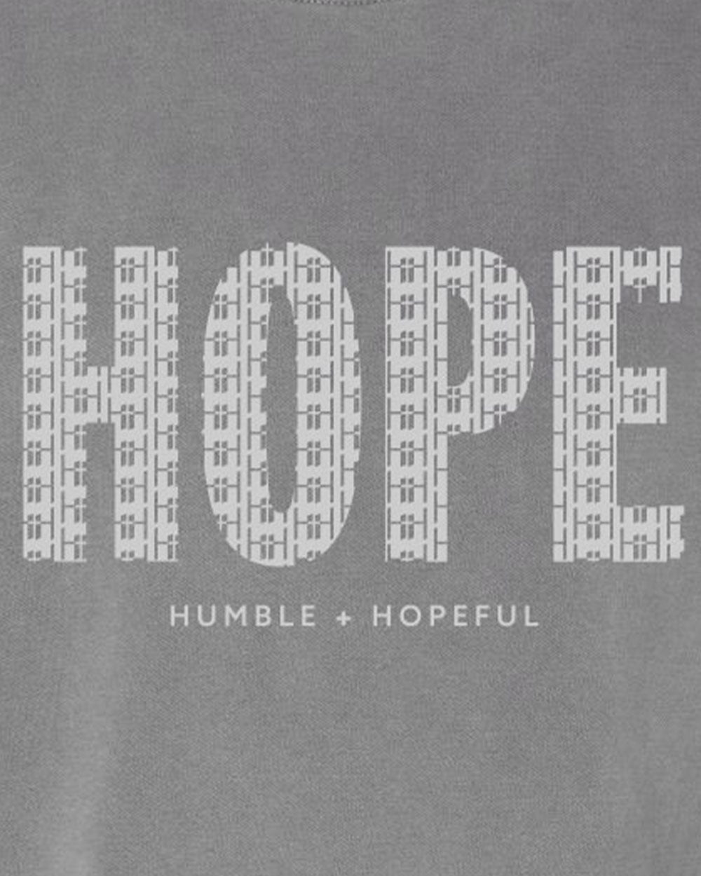 Close-up of a gray fabric surface with a large white 'HOPE' wordmark made of small cross-like shapes, and smaller text below reading 'HUMBLE + HOPEFUL'.