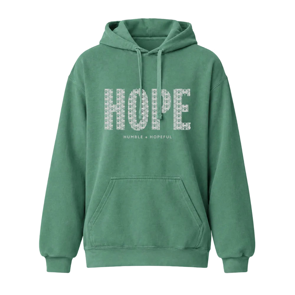 A green pullover hoodie with a front kangaroo pocket and drawstrings, featuring the word 'HOPE' printed across the chest in a white geometric pattern, with the text 'HUMBLE + HOPEFUL' underneath.