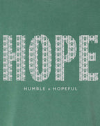 A close-up of a green garment featuring a white graphic print that reads 'HOPE' in a patterned font, with 'HUMBLE + HOPEFUL' printed underneath.