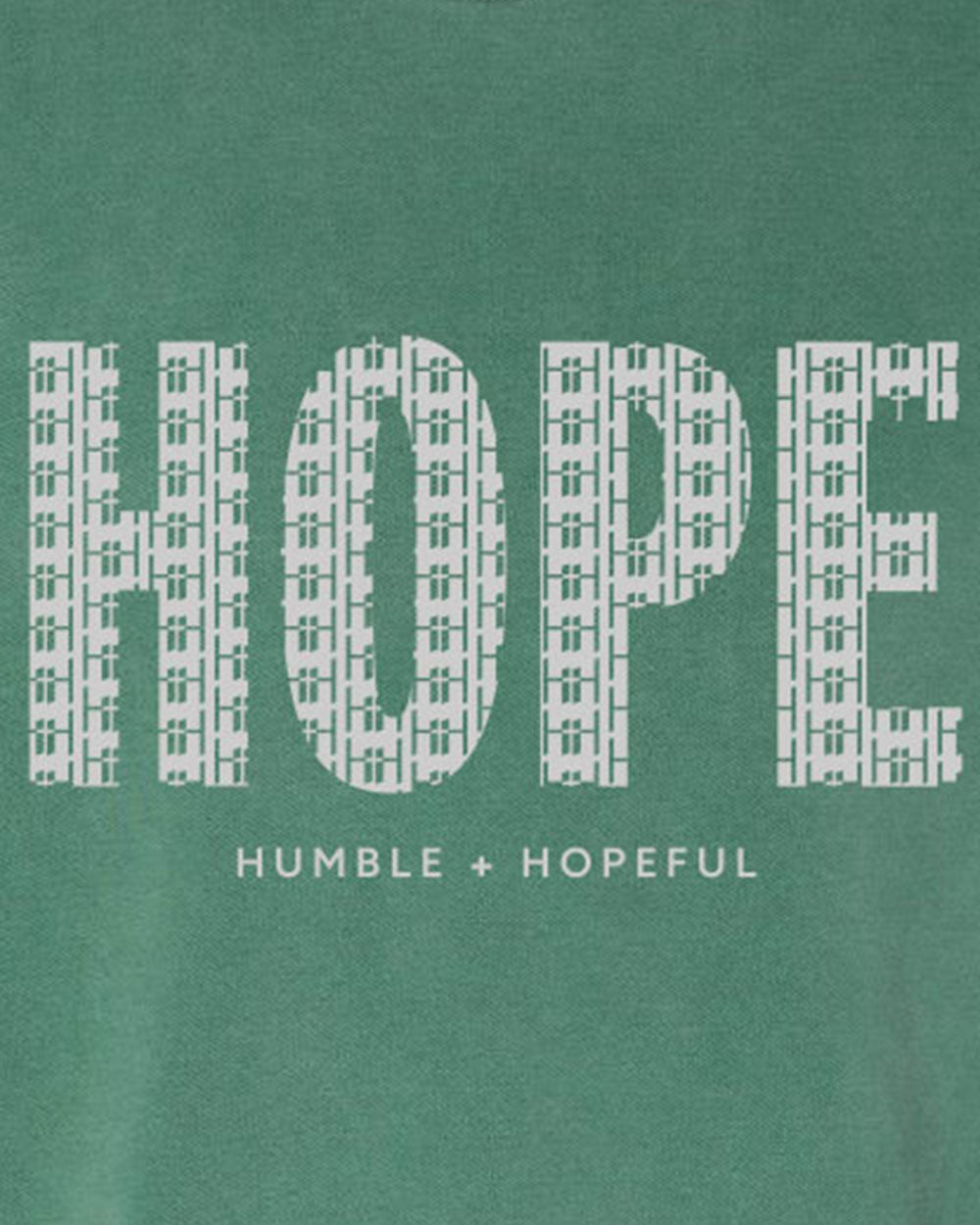 A close-up of a green garment featuring a white graphic print that reads 'HOPE' in a patterned font, with 'HUMBLE + HOPEFUL' printed underneath.