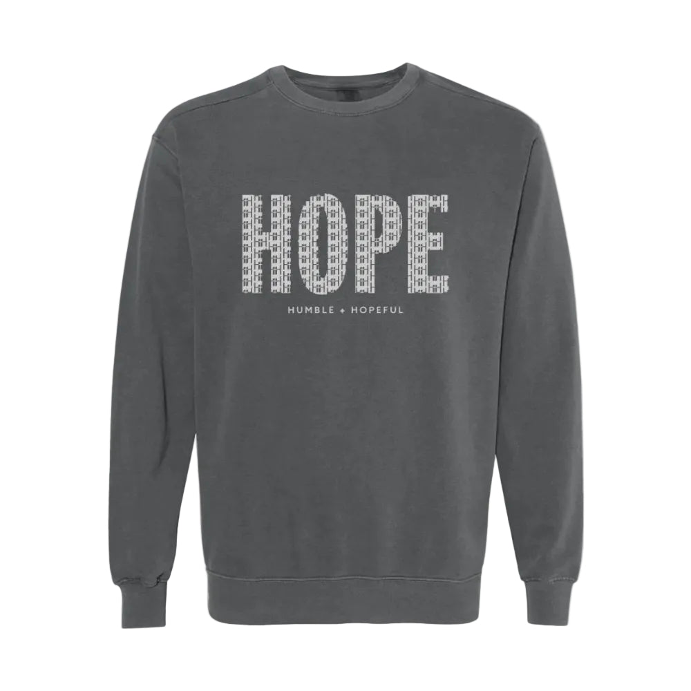 A dark gray crewneck sweatshirt featuring the word 'HOPE' printed across the chest in a white patterned font, with 'HUMBLE + HOPEFUL' written underneath in smaller text.
