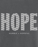A close-up of a dark gray graphic print featuring the word 'HOPE' in large white letters filled with a repeating cross pattern, with the phrase 'HUMBLE + HOPEFUL' printed below in smaller white text.