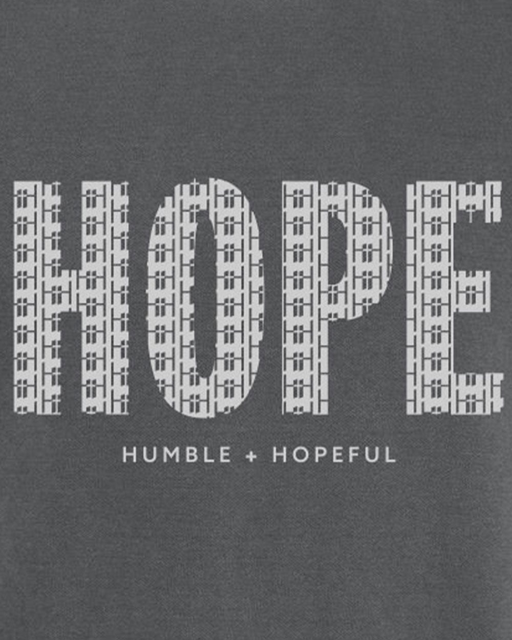 A close-up of a dark gray graphic print featuring the word 'HOPE' in large white letters filled with a repeating cross pattern, with the phrase 'HUMBLE + HOPEFUL' printed below in smaller white text.