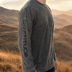 A man wearing a pepper gray long-sleeve t-shirt with 'GRATITUDE' printed vertically on the sleeve, standing in a scenic outdoor mountain landscape at sunset.