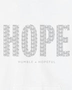 A close-up of a white garment featuring a large gray graphic that spells 'HOPE' in a patterned font, with the phrase 'HUMBLE + HOPEFUL' printed underneath.