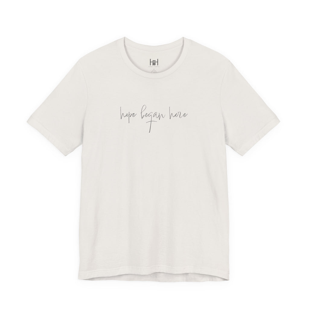 An off-white short-sleeve crewneck t-shirt with the phrase 'hope began here' printed in a thin gray cursive font across the chest. The shirt is displayed flat against a solid white background.