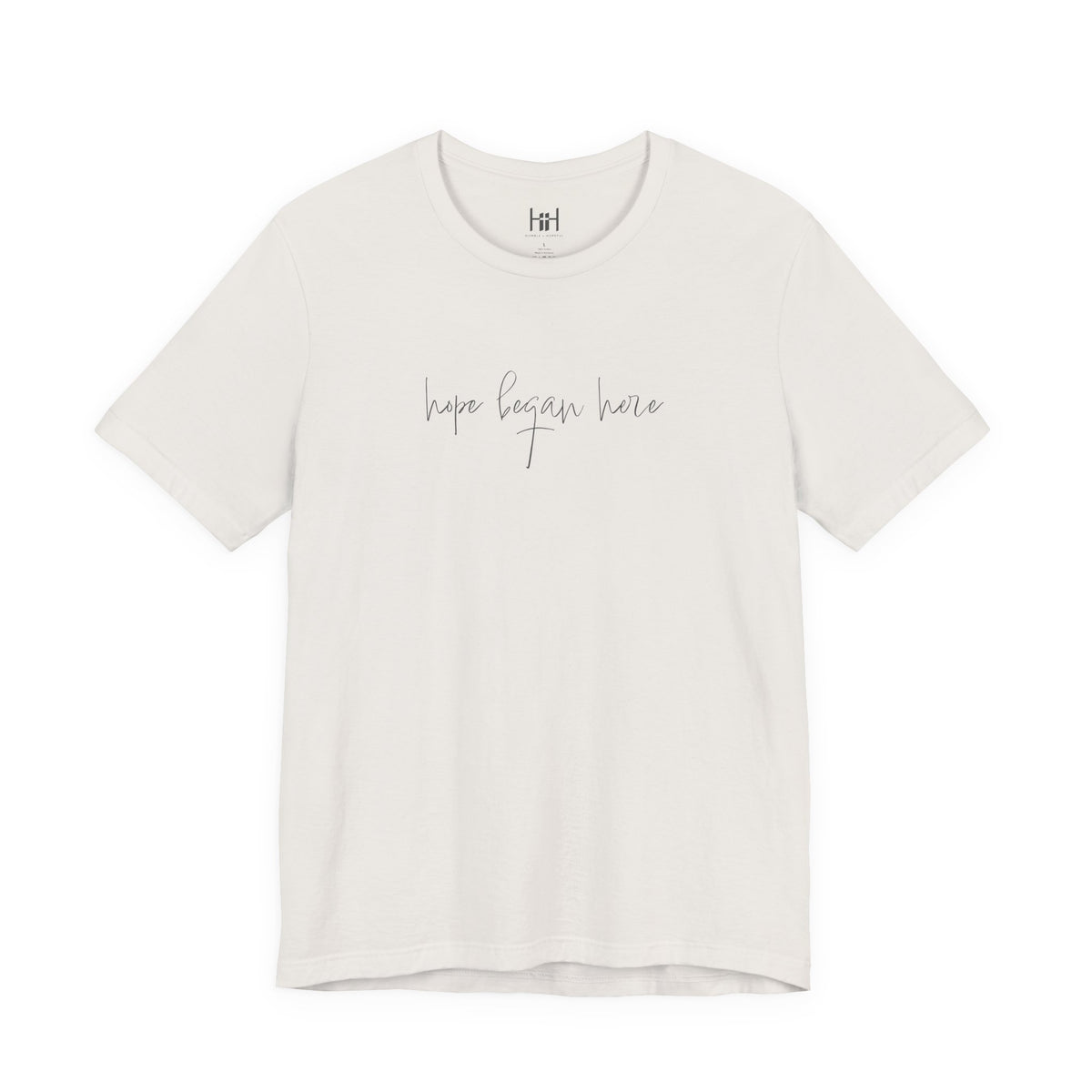 An off-white short-sleeve crewneck t-shirt with the phrase 'hope began here' printed in a thin gray cursive font across the chest. The shirt is displayed flat against a solid white background.